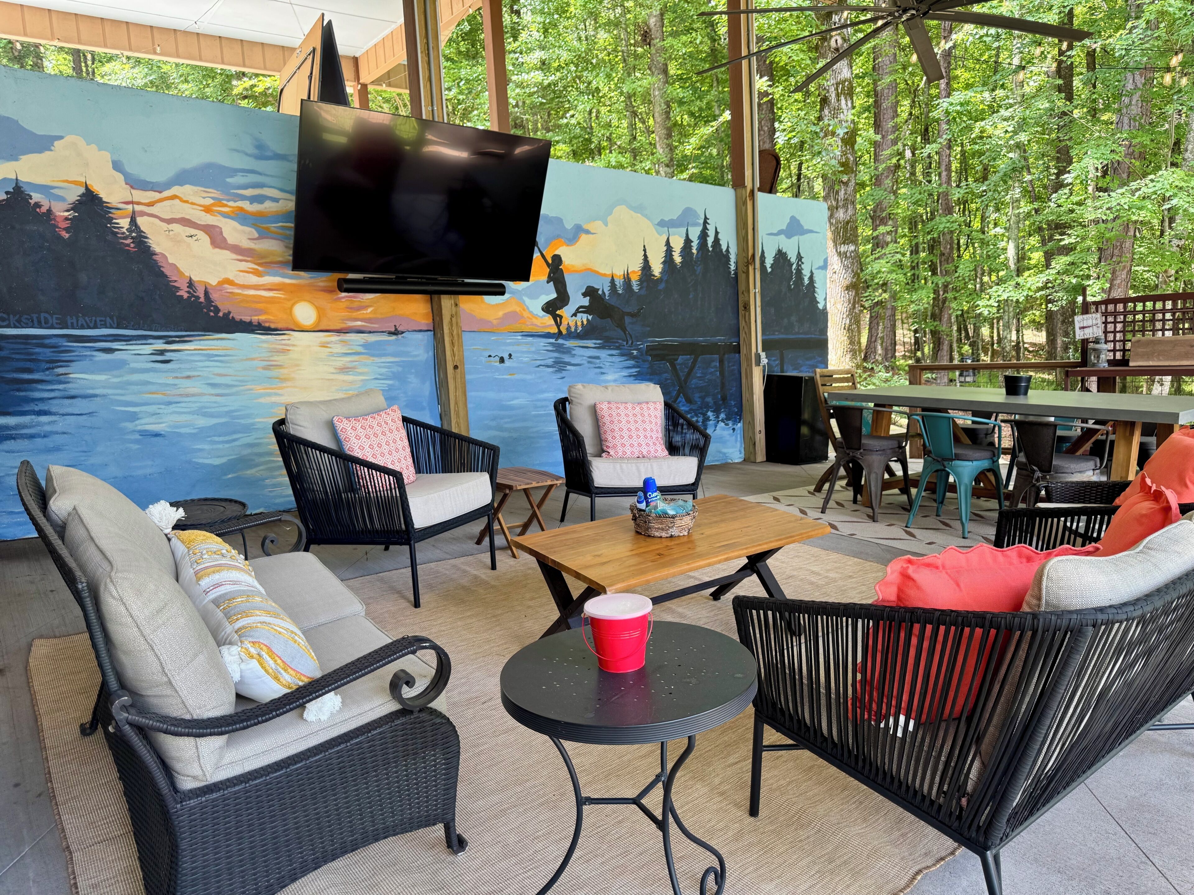 An outdoor seating area with wicker furniture, a wooden table, and a mural of a sunset over water.