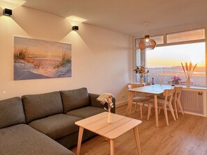 A modern living room with a sofa, a dining table with chairs, a painting, and a view through a large window. - Sea view in the vacation park - House G (Heiligenhafen)