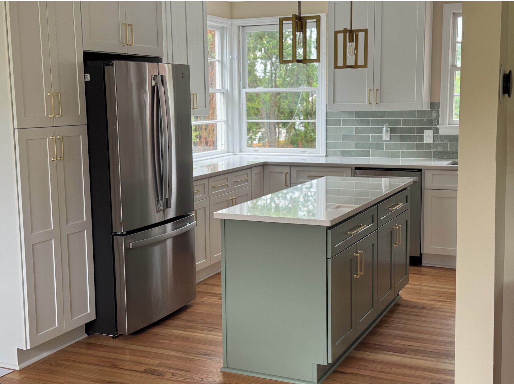 A modern kitchen with a stainless steel refrigerator, light-colored cabinets, a large island, and a window with a view of greenery.