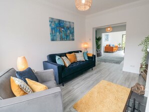 A modern living room with a dark blue sofa, a grey armchair, and a coffee table. There is a painting on the wall, a floor lamp, and a doorway leading to another room. - Riverside House (Scarborough)
