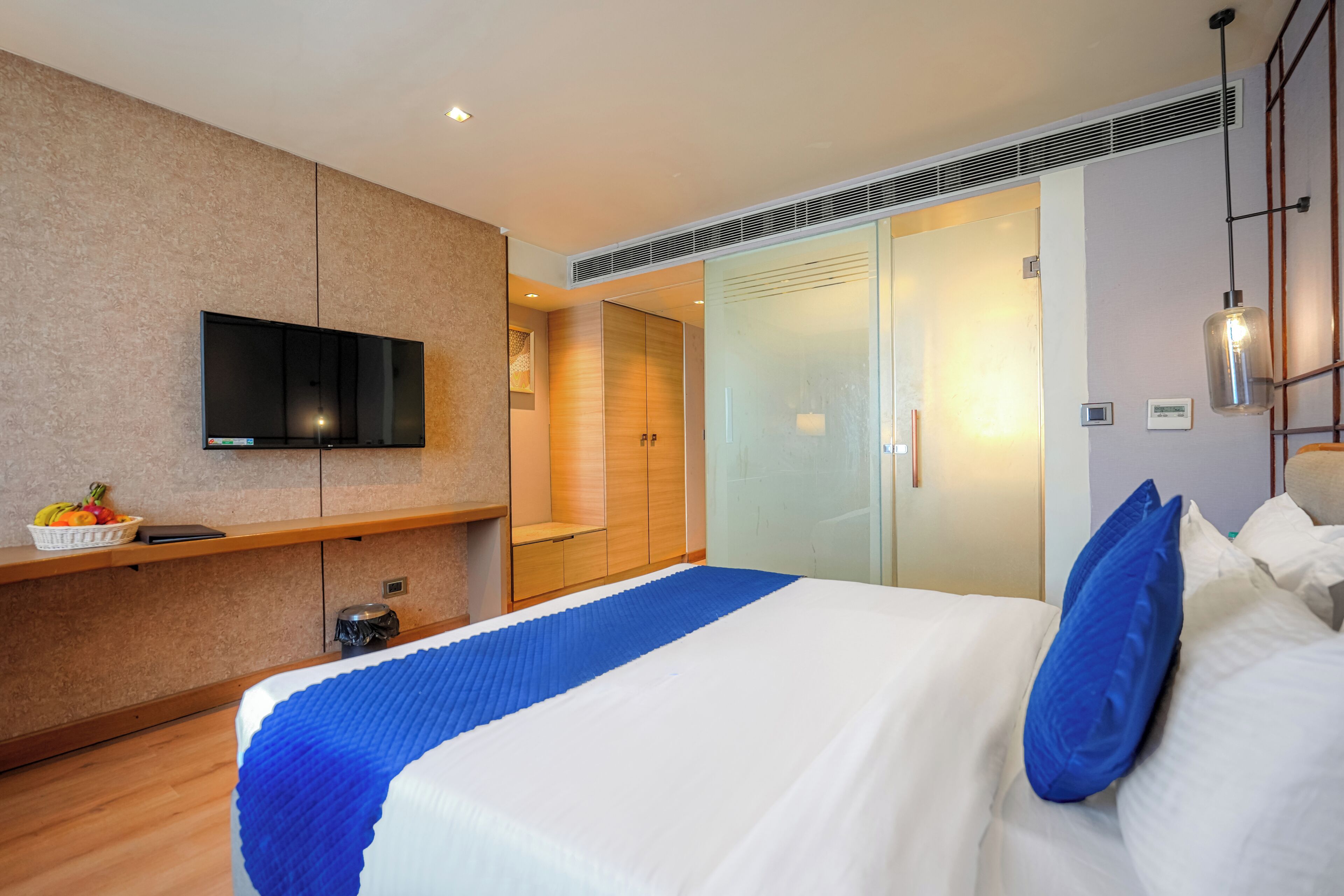 Une chambre d’hôtel moderne équipée d’un lit, d’un téléviseur à écran plat, d’une armoire et d’un petit coin salon agrémenté d’un panier de fruits.