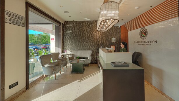 A reception area with a modern design, including a reception desk, seating area with a sofa and chairs, a large chandelier, and a view of the parking lot through the glass door. - Ramee Collection Airport Road Mohali (Mohali)