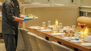 A chef in a uniform holding a plate of food, a dining table with various dishes, and a wall with framed artwork. - Ramee Collection Airport Road Mohali (Mohali)