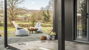 A modern outdoor seating area with a wooden deck, two bean bags, and a metal-framed door. - Grove Cottage (Kington)