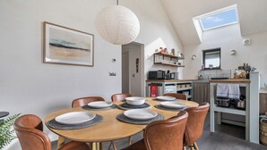 A dining area with a round table, brown chairs, a hanging light fixture, a kitchen with a window, and a framed picture on the wall. - Grove Cottage (Kington)