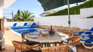 An outdoor pool area with lounge chairs, a dining table with chairs, and a large umbrella. - Casa Joshollie 3 bedroom villa (Carvoeiro)
