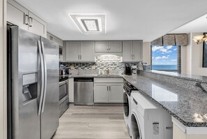 A modern kitchen with stainless steel appliances, grey cabinetry, a granite countertop, and a view of the sky through the window. - Waterpointe II 701 l Oceanfront l 3BR l Pools (North Myrtle Beach)