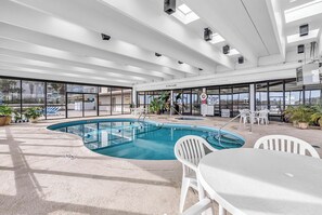 An indoor swimming pool with clear signage, a sunlit patio area with seating, and a view of the surrounding buildings. - Waterpointe II 701 l Oceanfront l 3BR l Pools (North Myrtle Beach)