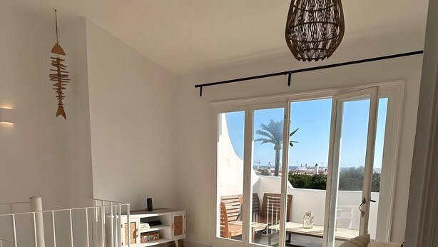 Amplia habitación con piso de madera, barandal blanco, ventanal con vistas y lámpara colgante.