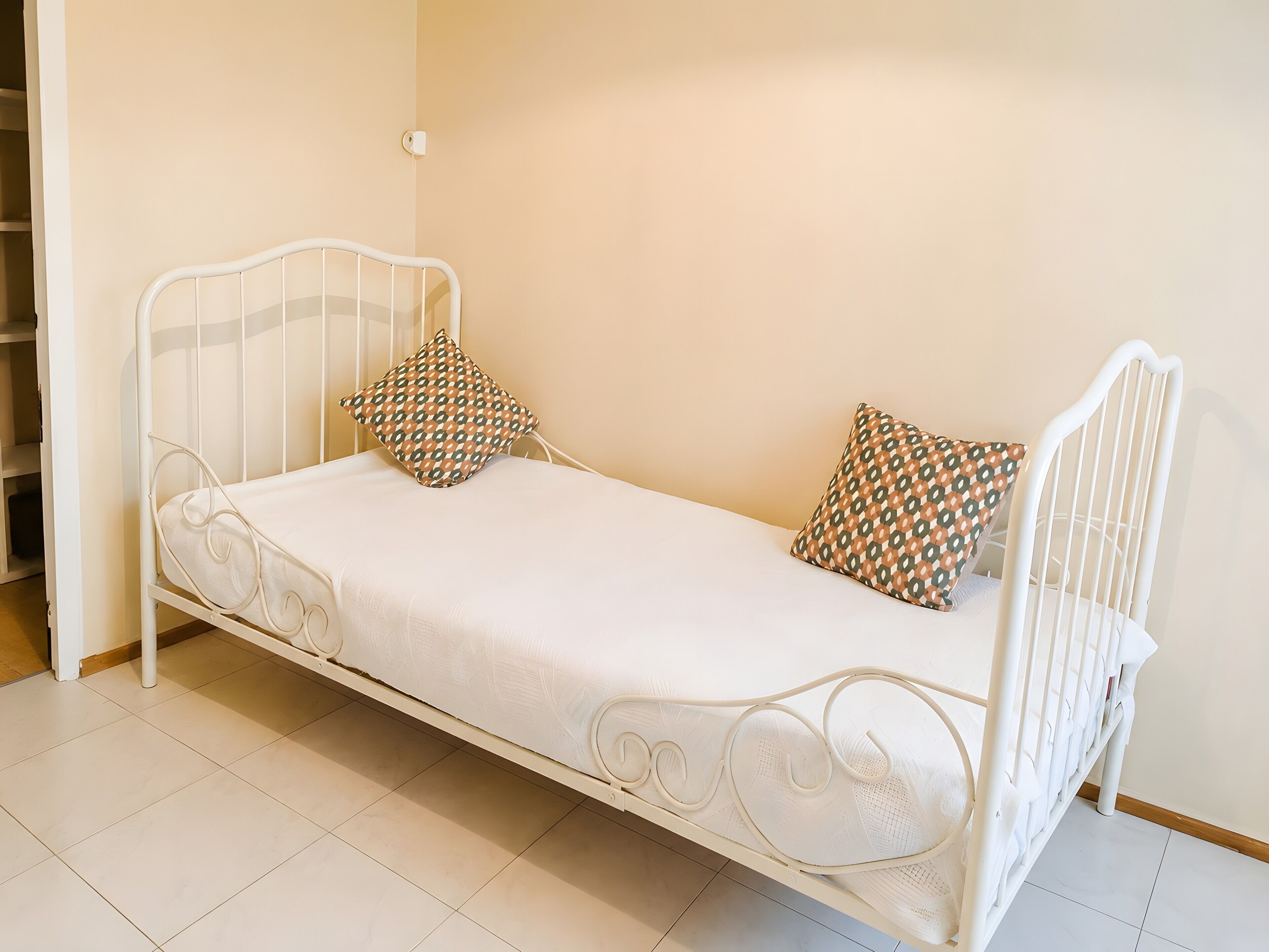 A white metal bed with two patterned pillows, a white mattress, and a plain wall.