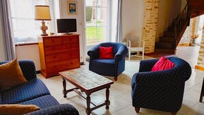 Un salon avec une commode en bois, un téléviseur, deux fauteuils bleus avec des coussins rouges, une petite table et un escalier en arrière-plan.