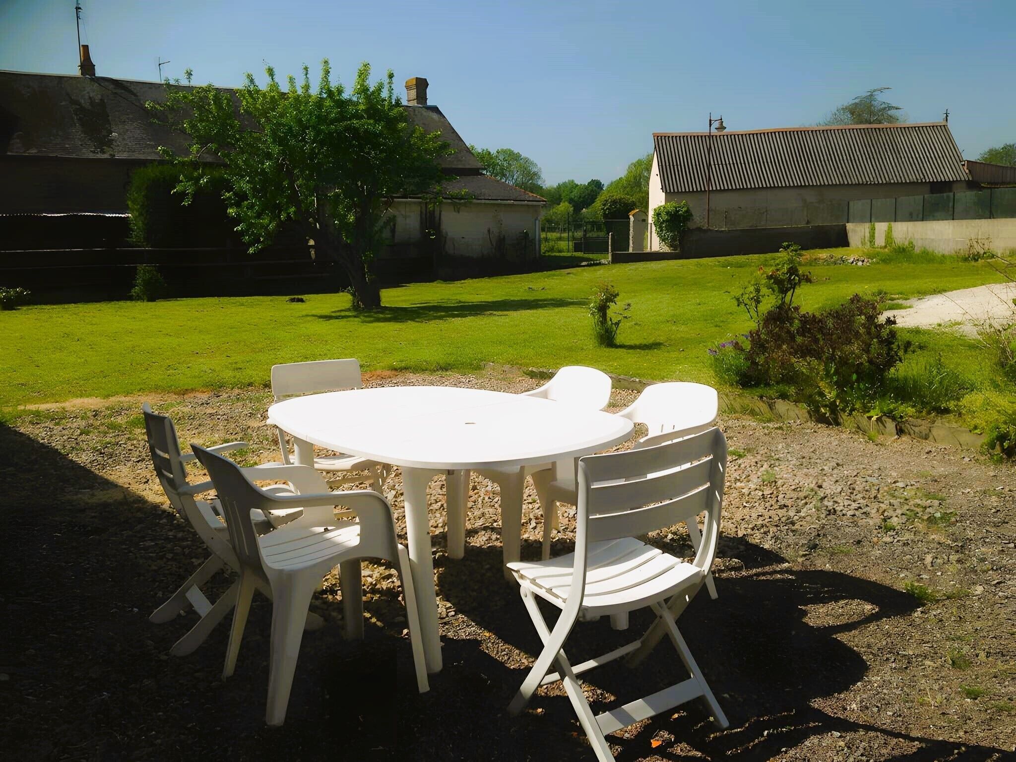 A round outdoor table with four chairs, surrounded by gravel and grass, with buildings in the background.