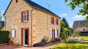 Une maison en pierre avec des volets rouges, une porte d’entrée et un petit jardin agrémenté d’une table et de chaises.