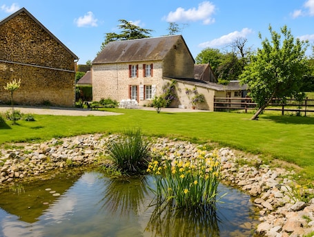 A stone building with a pond, surrounded by a well-maintained lawn and a wooden fence.. Country House with Private Terrace, Garden, and Wi-Fi in Sainte-Scolasse-sur-Sarthe