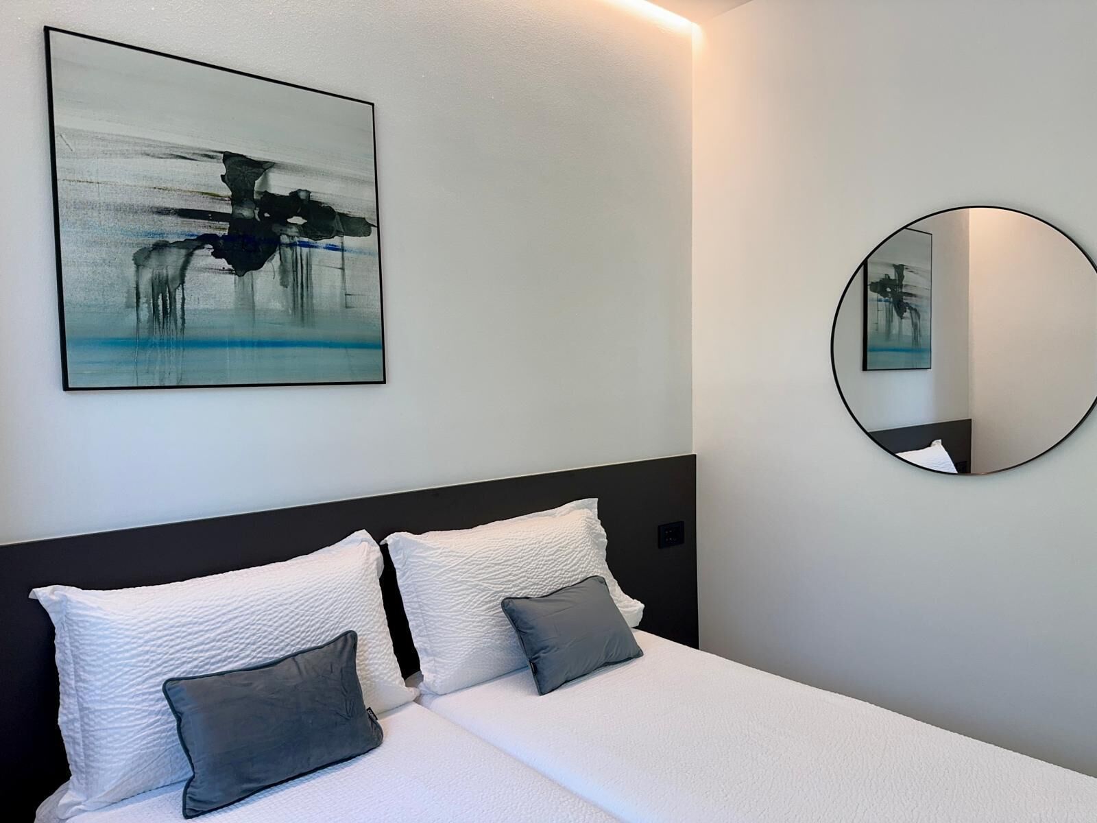 Een moderne slaapkamer met een bed, kussens, een ronde spiegel en een schilderij aan de muur.