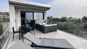 A modern outdoor balcony with a glass balustrade, a seating area with a sofa and chairs, and a hot tub. - Aria Art Suite   Lake View Terrace and Private Hot Tub (BARDOLINO)