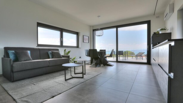 A modern living room with a grey sofa, a small round table, and a view of the outdoors through large sliding doors. - Aria Art Suite   Lake View Terrace and Private Hot Tub (BARDOLINO)
