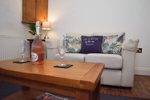 Ein Wohnzimmer mit Couch, tropischen Kissen, einem Holztisch, einer Weinflasche und zwei Weingläsern sowie einer Lampe.