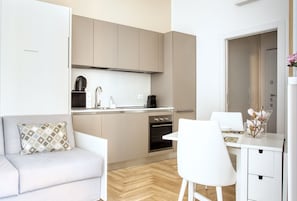 Eine kompakte Küche mit eingebautem Backofen, ein Essbereich mit weißem Tisch und Stuhl sowie eine Couch mit gemustertem Kissen.