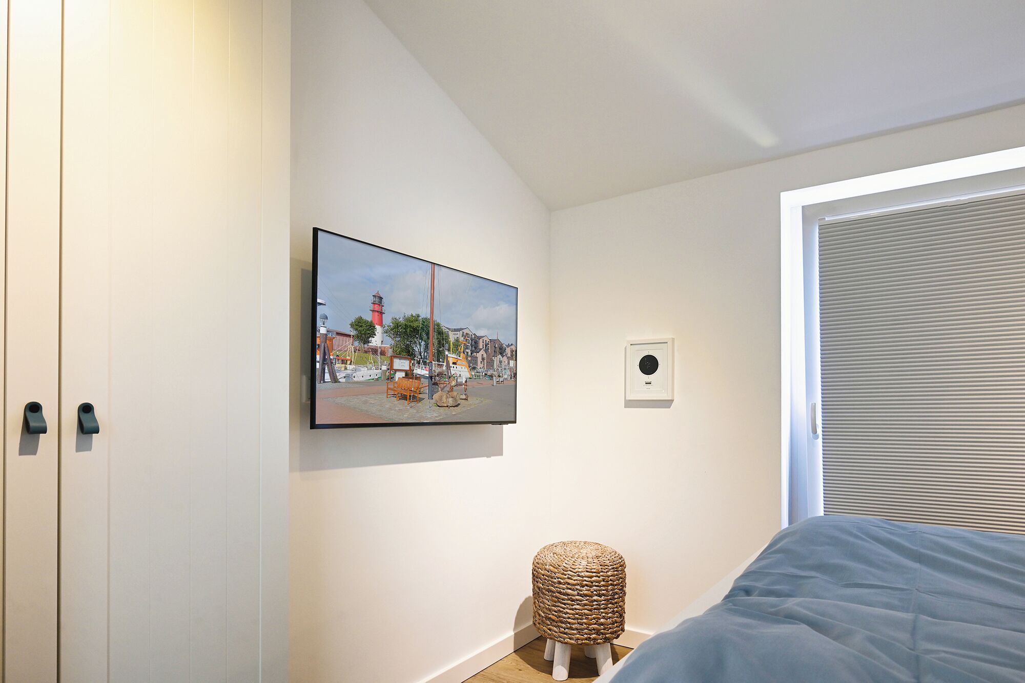 A bedroom with a bed, a wall-mounted picture, a small stool, and a window with blinds.