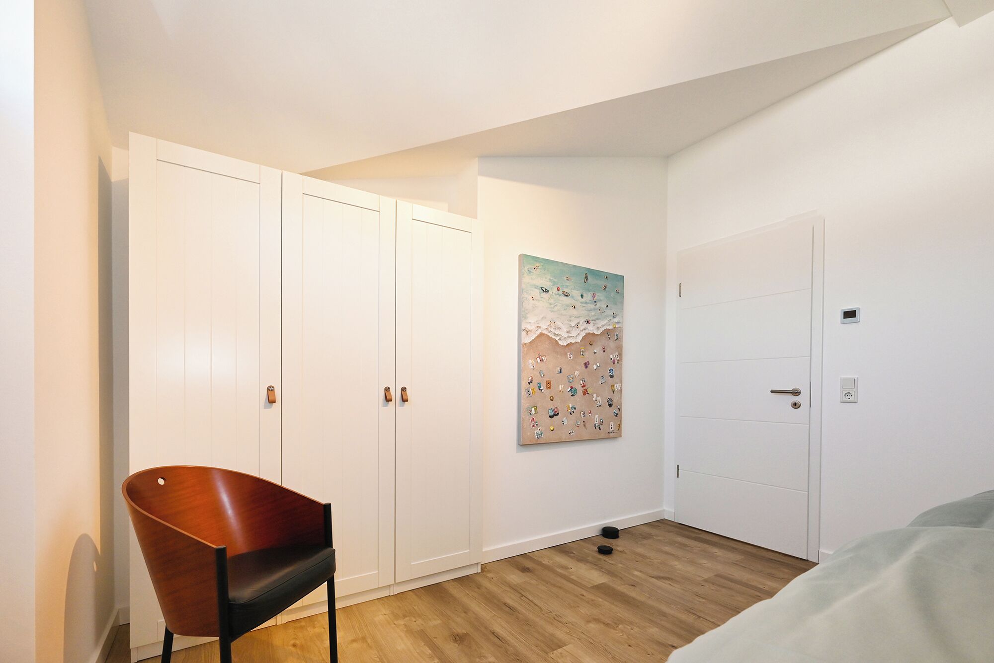 Ein Schlafzimmer mit Bett, einem Holzstuhl, einem Kleiderschrank, einer Tür und einem an der Wand hängenden Kunstwerk.