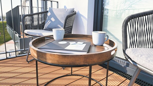 Un balcon avec une table ronde en bois, deux tasses blanches, un menu et une paire de chaises noires avec des coussins.