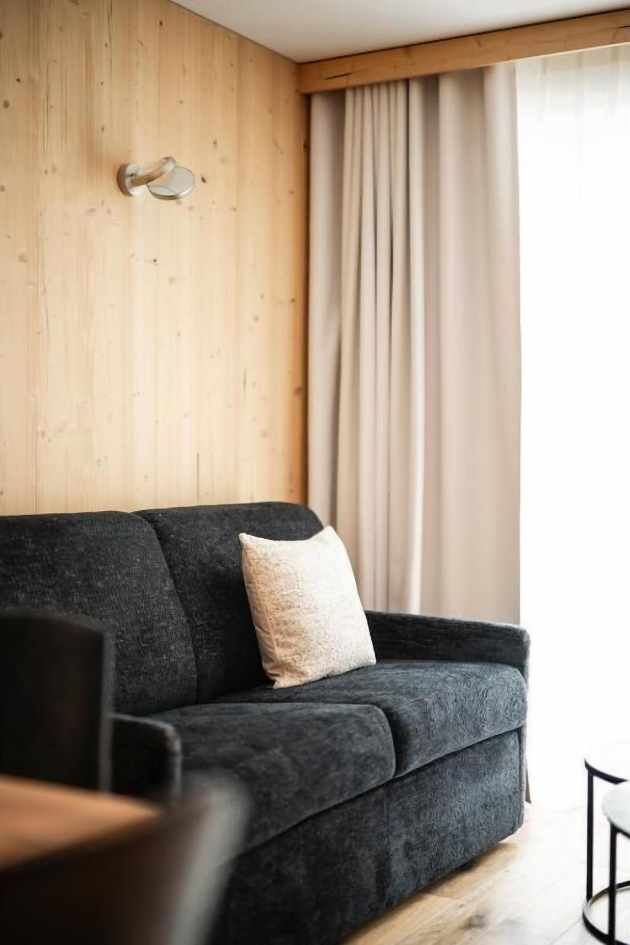 Una sala de estar moderna con un sofá gris oscuro, un cojín de color claro, una pared de madera y un amplio ventanal con cortinas.