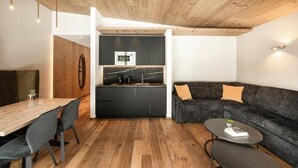 A modern interior with a wooden ceiling, a dining area with a wooden table and chairs, a black kitchen with a marble backsplash, and a dark gray sofa with cushions. - Hotel La Truga (Selva di Val Gardena)
