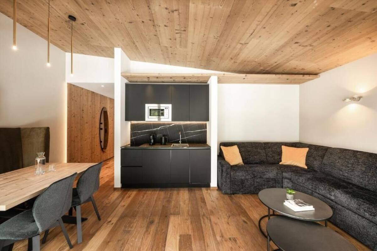 Een modern interieur met een houten plafond, een eethoek met een houten tafel en stoelen, een zwarte keuken met een marmeren spatwand, en een donkergrijze bank met kussens.