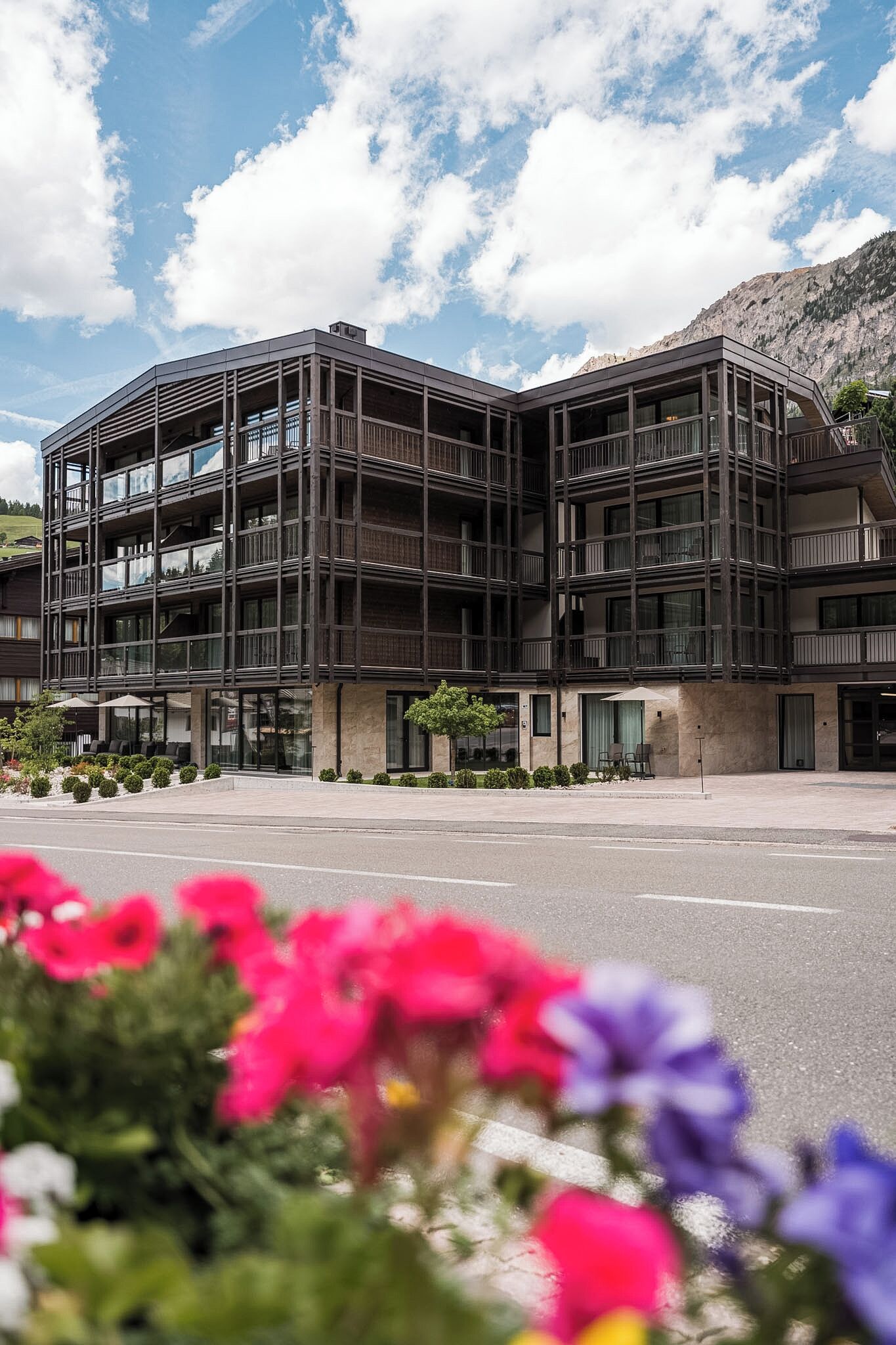 A modern multi-story building with balconies, large windows, and a landscaped area with flowers.