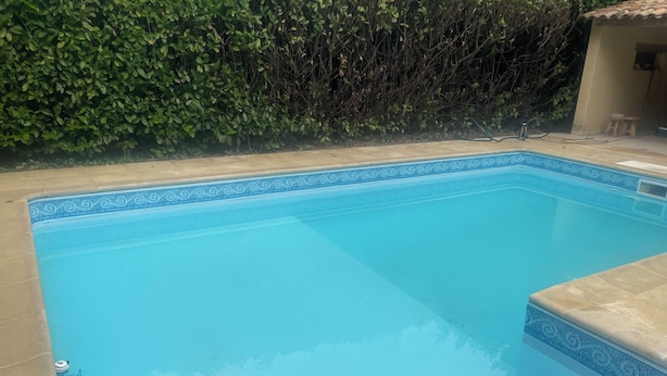 Une piscine rectangulaire dont les bords sont recouverts de carreaux, entourée d’une terrasse en pierre et d’une haie.