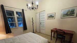 Une chambre à coucher comprenant un lit, un bureau, une chaise, un porte-manteau, une porte, une fenêtre avec des rideaux et un lustre.