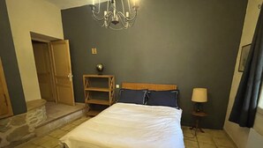Une chambre à coucher comprenant un lit, une tête de lit en bois, une table de chevet, une lampe, un lustre et une porte donnant sur une autre pièce.