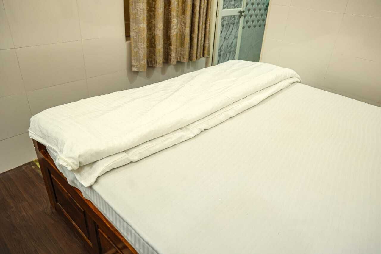 A neatly made bed with a white mattress and a white blanket, a wooden headboard, and a curtain partially drawn to the side.