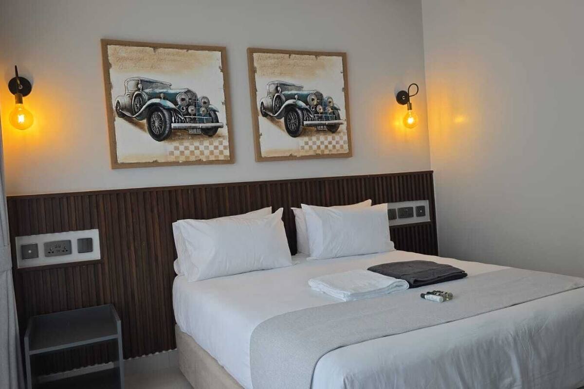 Habitación de hotel con una cama, dos lámparas fijadas a la pared y dos cuadros antiguos de automóviles.