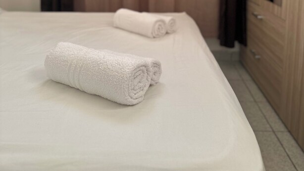 Een hotelkamer met een bed, twee opgerolde witte handdoeken, een houten garderobe en een raam met gordijnen.