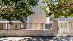 A paved courtyard with a metal fence, a building with vertical siding, and a tree providing shade. - Holiday Home 'Casa Dafne in Otranto 6 Guests' with Balcony, Wi-Fi and Air Conditioning (Otranto)