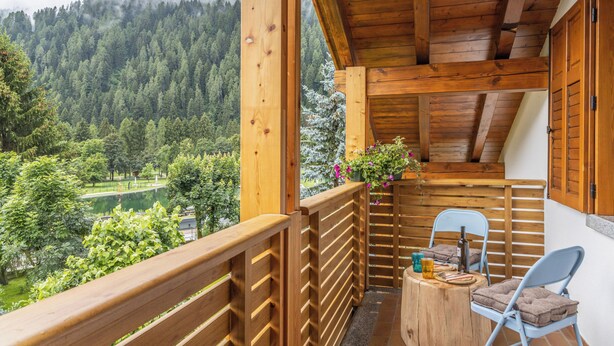 Un balcón de madera con vistas a un valle boscoso, una mesa pequeña con sillas y una botella.