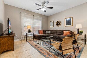 A living room with a sofa, coffee table, and chairs, a television, and a ceiling fan. - 4BR Townhome•Pvt pool•Near theme parks (Kissimmee)
