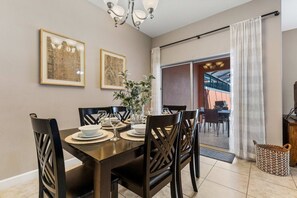 Un coin repas avec une table en bois dressée pour quatre personnes, entourée de chaises, avec un lustre au-dessus et une grande fenêtre agrémentée de rideaux.