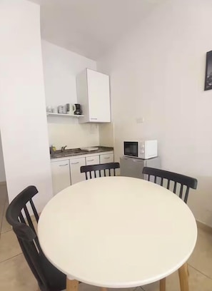 Cocina compacta con armarios blancos, microondas y una pequeña encimera. Hay una mesa redonda con cuatro sillas.