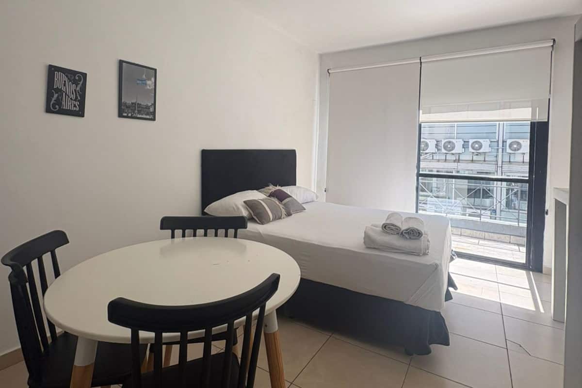 Una habitación de hotel con una cama, una mesa redonda con sillas, una ventana que ofrece vistas a edificios y dos cuadros colgados en la pared.