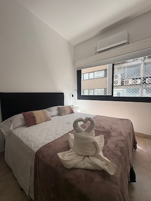 Quarto de hotel com cama, janela com vista para edifícios e unidade de ar condicionado na parede.