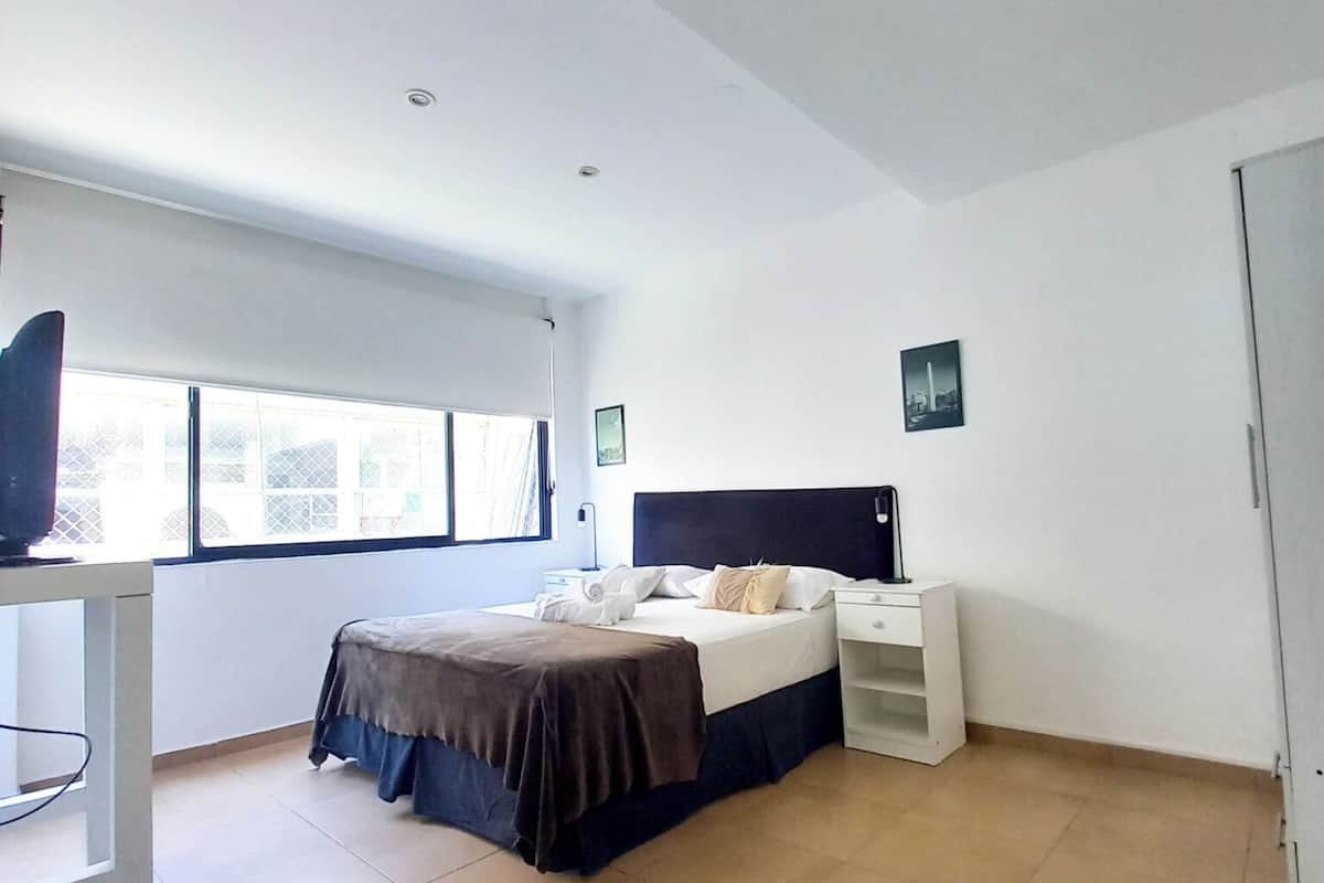 Un dormitorio con cama, mesitas de noche, un escritorio y una ventana.