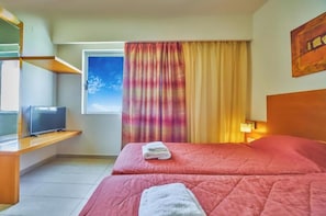 Una habitación de hotel con cama, televisor sobre un mueble, ventana con cortinas y un cuadro en la pared.
