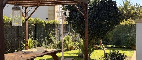 Una mesa de picnic de madera bajo una pérgola, con un jardín al fondo.