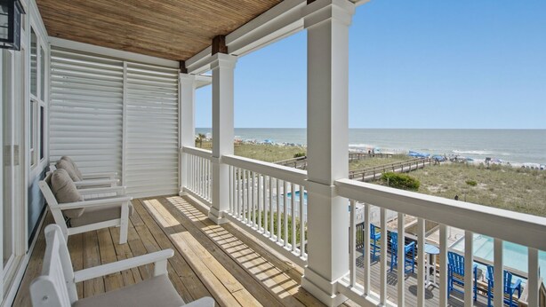 Une terrasse en bois avec des rampes blanches, une piscine et une vue sur la plage et l’océan.