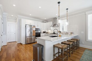 A modern kitchen with white cabinetry, stainless steel appliances, a central island with stools, and pendant lighting. - Kure Surf Shack - 5 bed, oceanfront with large pool and elevator (Kure Beach)