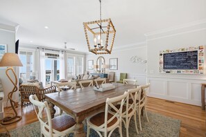 A dining area with a wooden table, white chairs, a woven floor lamp, and a decorative hanging light fixture. - Kure Surf Shack - 5 bed, oceanfront with large pool and elevator (Kure Beach)