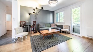 Een moderne woonkamer met een bank, salontafel en stoelen. Er is een eettafel met stoelen, een groot raam en een hanglamp.